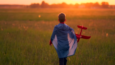 Der-Junge-Im-Kostüm-Eines-Superhelden-Läuft-In-Einem-Roten-Mantel-Und-Lacht-Bei-Sonnenuntergang-Auf-Einer-Sommerwiese-Und-Stellt-Sich-Vor,-Er-Sei-Der-Pilot-Des-Flugzeugs-Und-Spiele-Mit-Einem-Modellflugzeug.