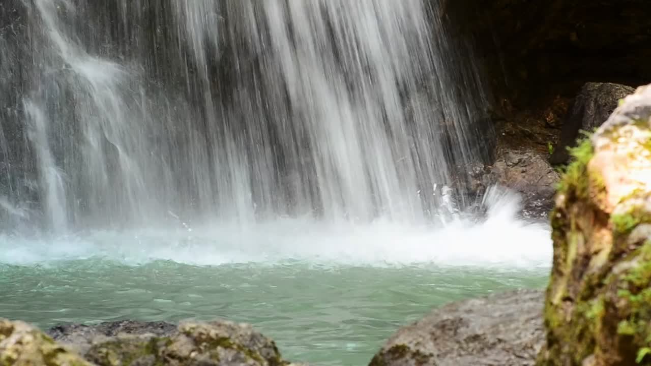 primer plano estacionario de agua chocando contra una cuenca turquesa detrás de grandes rocas cubiertas de musgo