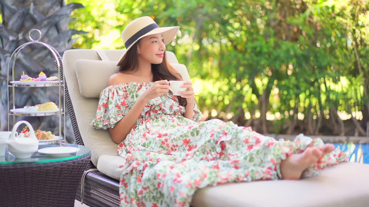 Woman in sundress with floral pattern and straw hat relaxing on chair beside pool with trays of fruit and pot of tea. Luxury resort relaxation and lifestyle concept. Asian lady having breakfast