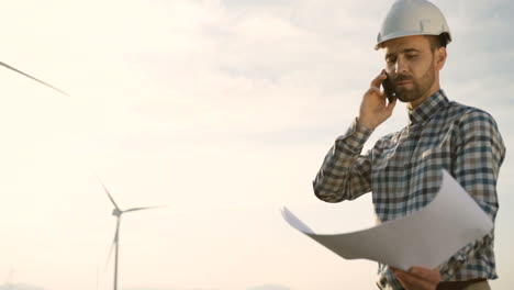 ingeniero caucásico con casco hablando por teléfono y viendo algunos planos en la estación eólica de energía renovable