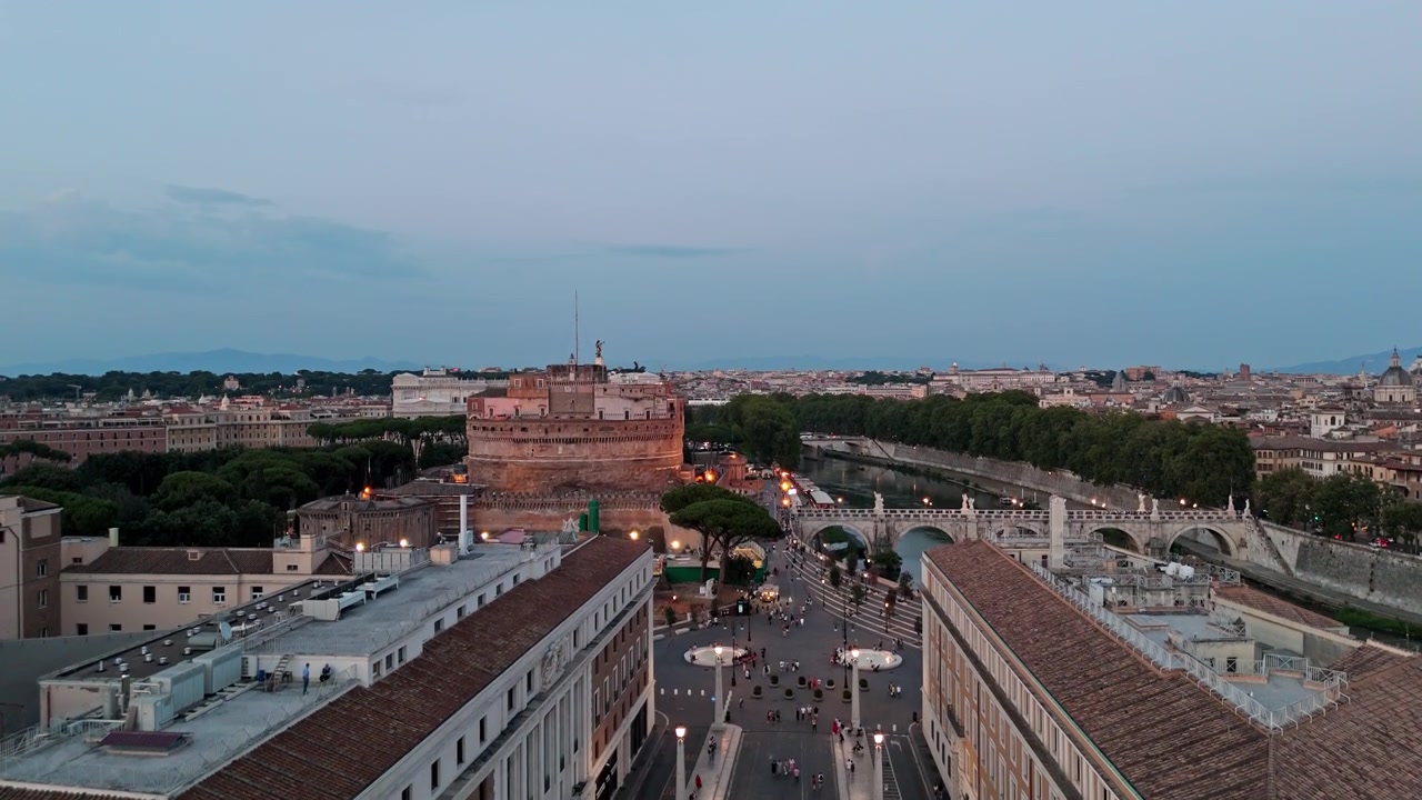Sorvolo serale con drone che scivola sopra Via della Conciliazione mentre il traffico e i pedoni si muovono verso Castel Sant'Angelo e il lungofiume.
