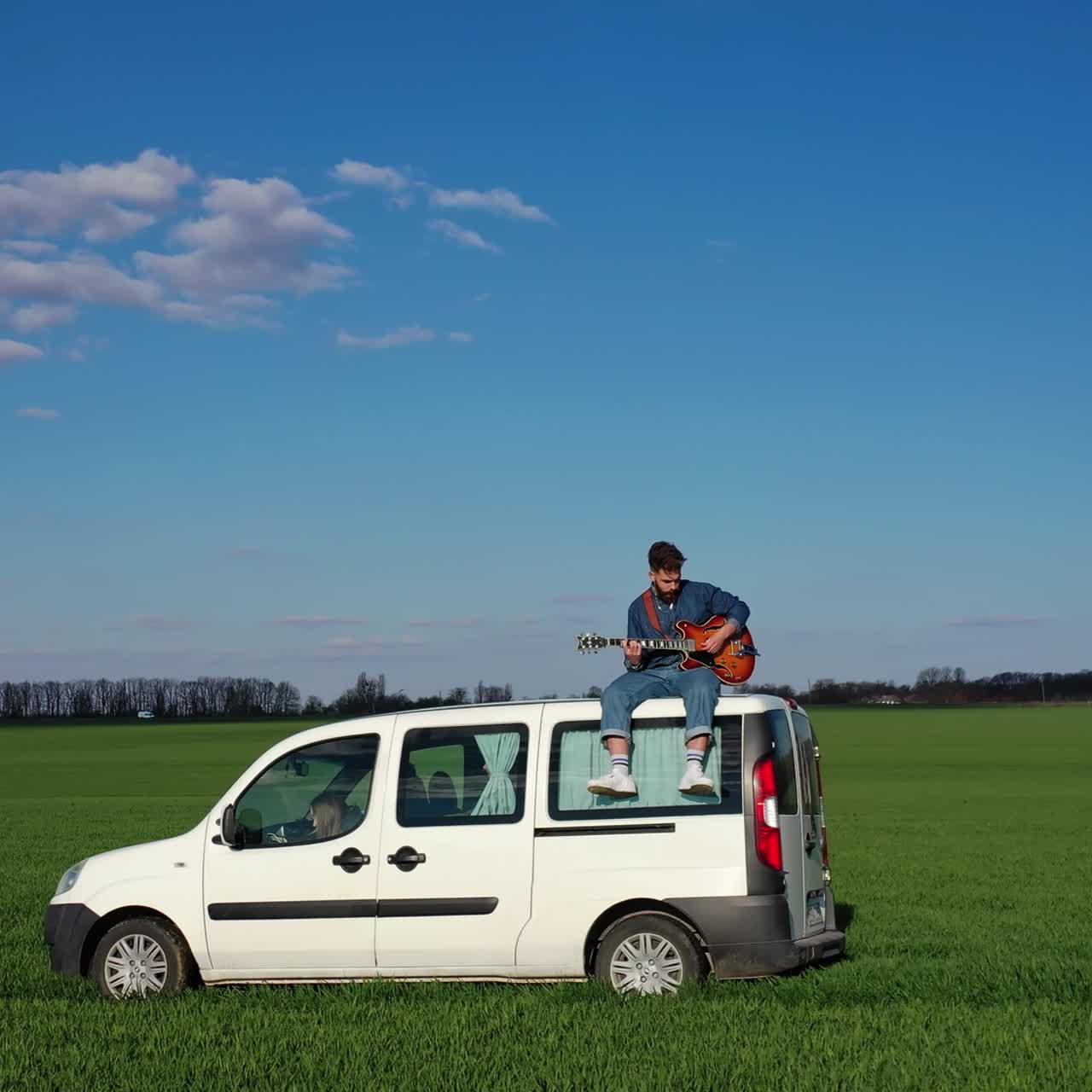 Young man with a guitar on a car moving on green field. Hipster guy likes extreme. Man playing the guitar while sitting on the top of a white car driving slowly in nature