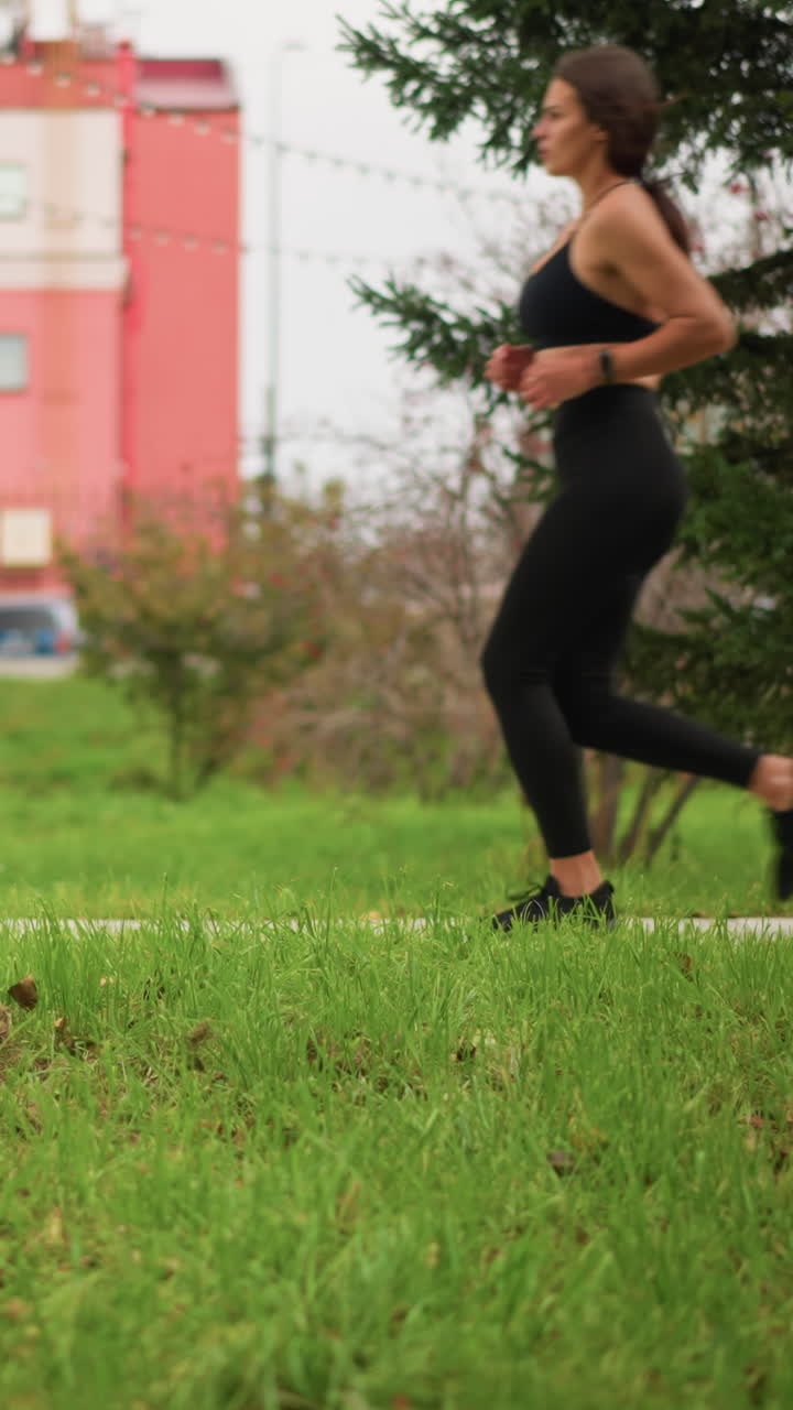 Background featuring grass, poles, and building with lady jogging on street path wearing black leggings, athletic woman in motion with focus on outdoor jogging in urban setting