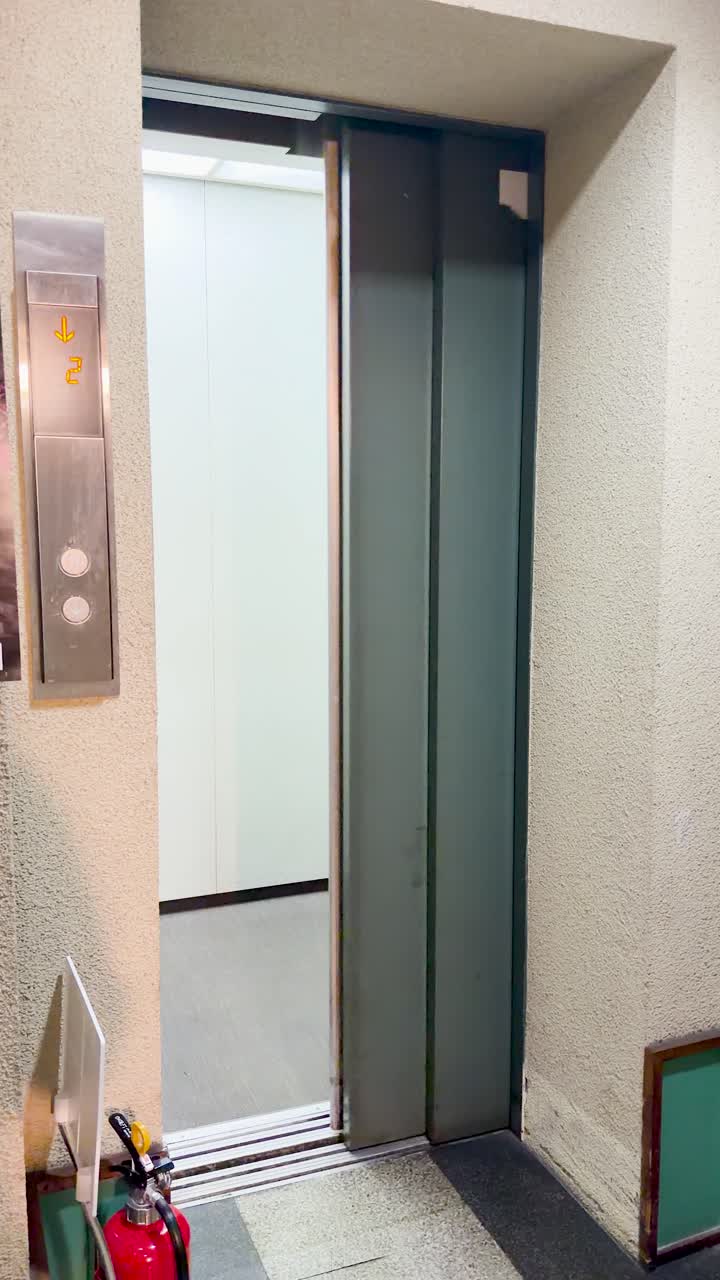Inquadratura statica di porte di ascensore grigie che si aprono per rivelare un interno di ascensore luminoso e vuoto.