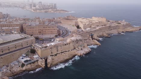 Drone-Elevándose-Sobre-El-Casco-Antiguo-De-Valletta-Malta-En-Un-Día-Brumoso-Con-Océano-Y-Olas