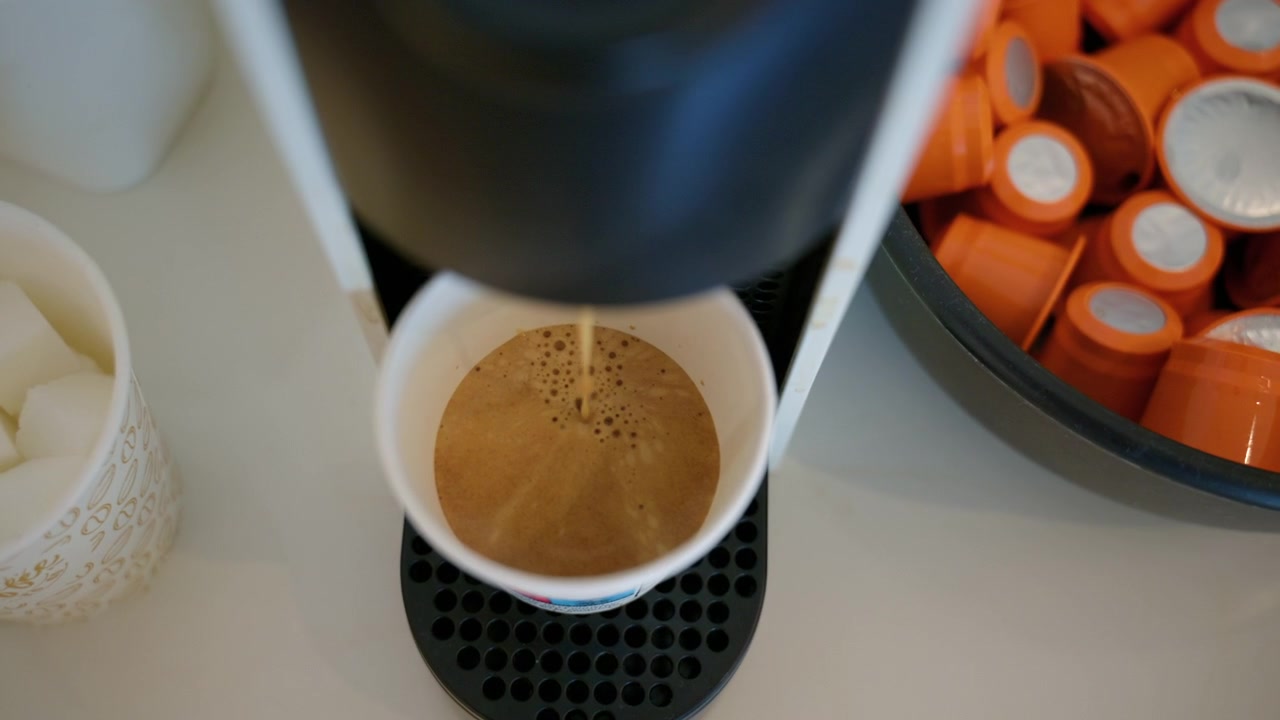 Coffee machine preparing a fresh espresso shot into a disposable cup, surrounded by coffee pods, sugar cubes, and wooden stirrers
