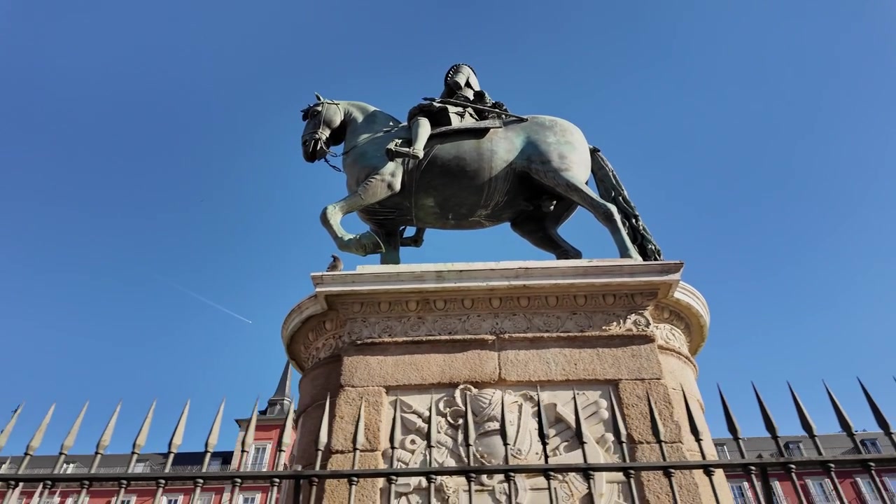 Una ripresa con slider a mano rivela la statua in bronzo di Filippo III in cima al suo piedistallo di pietra ornato nella Plaza Mayor di Madrid, incorniciata da architettura storica e una luce diurna vivida e senza nuvole