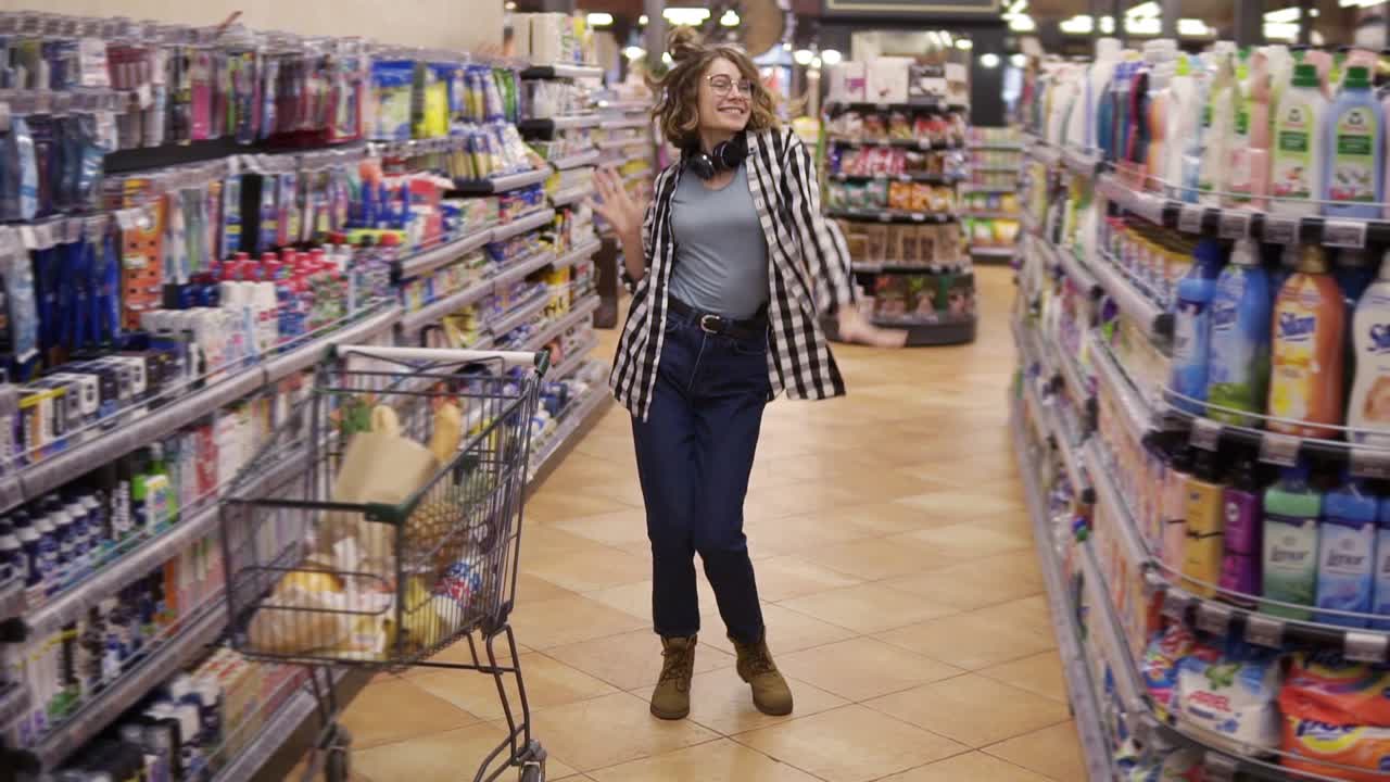 imágenes completas de una mujer joven en vaqueros y camisa a cuadros bailando de pie en el pasillo de la tienda de comestibles. emocionada con auriculares en el cuello mujer divirtiéndose, bailando en el supermercado. cámara lenta