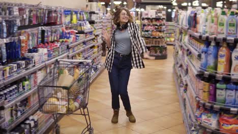 Imágenes-Completas-De-Una-Mujer-Joven-Con-Jeans-Y-Camisa-A-Cuadros-Bailando-Parada-En-El-Pasillo-De-Una-Tienda-De-Comestibles.-Emocionado-Con-Auriculares-En-El-Cuello-Mujer-Divirtiéndose,-Bailando-En-El-Supermercado.-Camara-Lenta