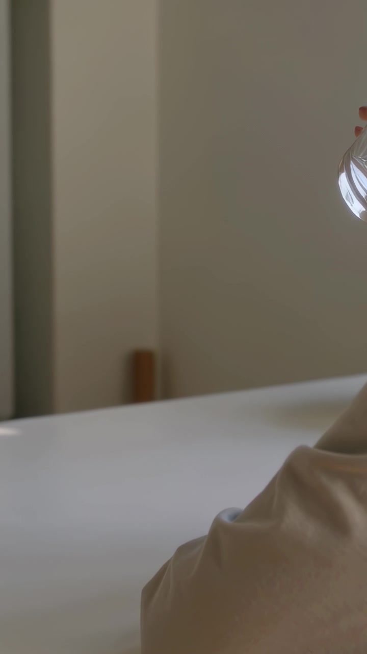 Vertical video: Tilting beige sleeve hand lifting glass to light at home white top peg, copy space