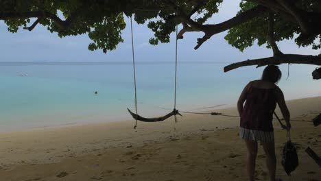 Eine-Frau-Stellt-Ihre-Tasche-Am-Strand-Ab,-Bevor-Sie-Auf-Eine-Holzschaukel-Springt