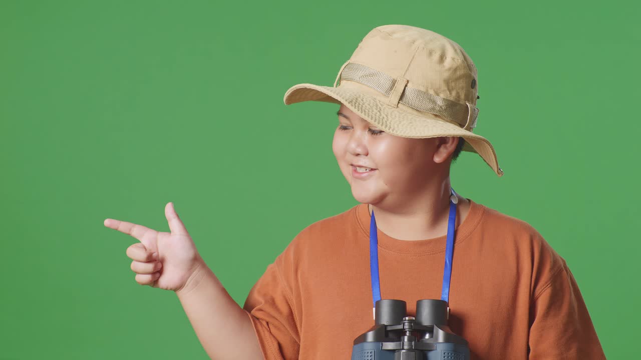 niño turista asiático con un sombrero y binoculares sonriendo y señalando hacia un lado mientras está de pie en el fondo de la pantalla verde. niño investigador examina algo, concepto de aventura de turismo de viaje, de cerca