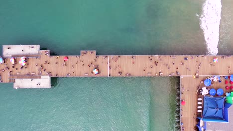 Santa-Monica-Pier-Von-Oben-Nach-Unten,-Vogelperspektive,-Drohne-Mit-Tosenden-Wellen-Und-Menschen,-Die-Auf-Der-Promenade-Laufen