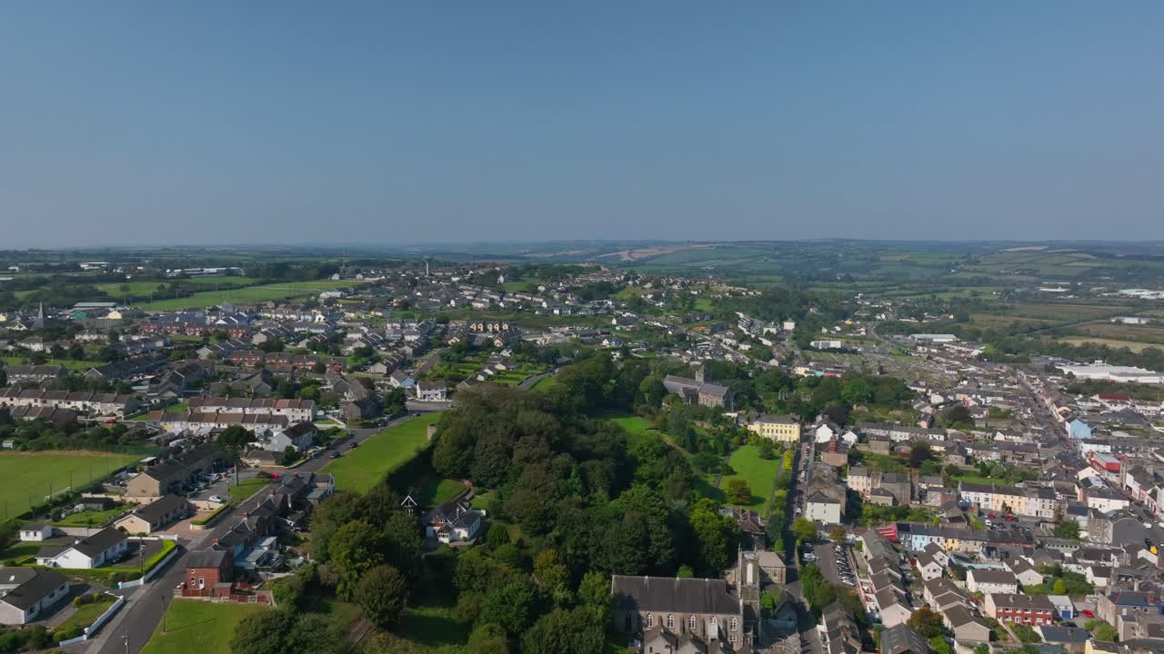 Youghal, County Cork, Ireland, September 2024. Drone pushes forward in panoramic high angle overview above town center with tree line separating traditional homes from new housing developments.