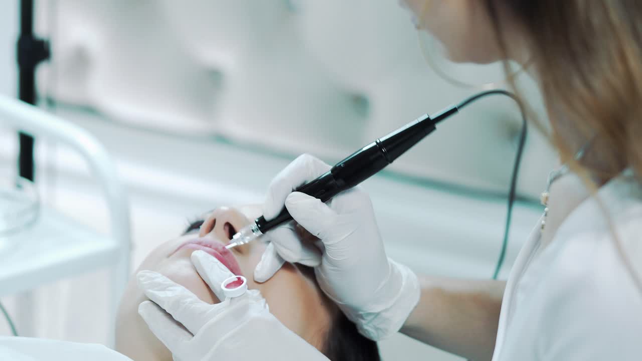 A beautician in white gloves does permanent lip makeup with red pigment. Beauty saloon