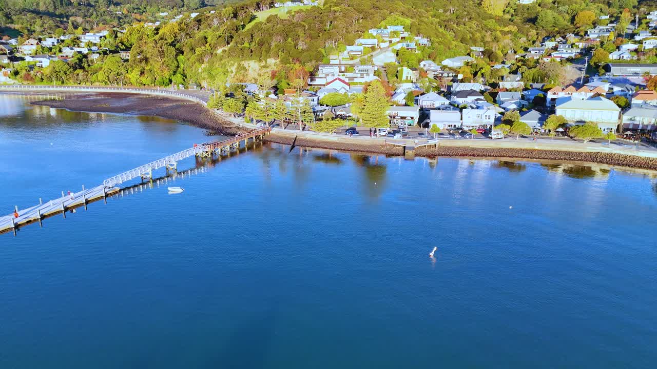 Drone footage captures Akaroa's scenic harbor, pier, and hillside homes under bright daylight, showcasing serene coastal beauty