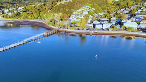 drone footage captures akaroa's scenic harbor, pier, and hillside homes under bright daylight, showcasing serene coastal beauty