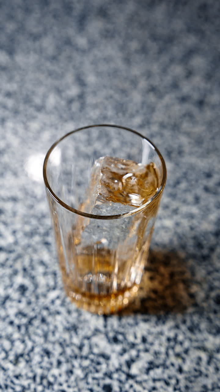 Thick yellow liquid syrup is poured into the glass with ice cube. Making a cocktail. Close up. Top view. Vertical video.