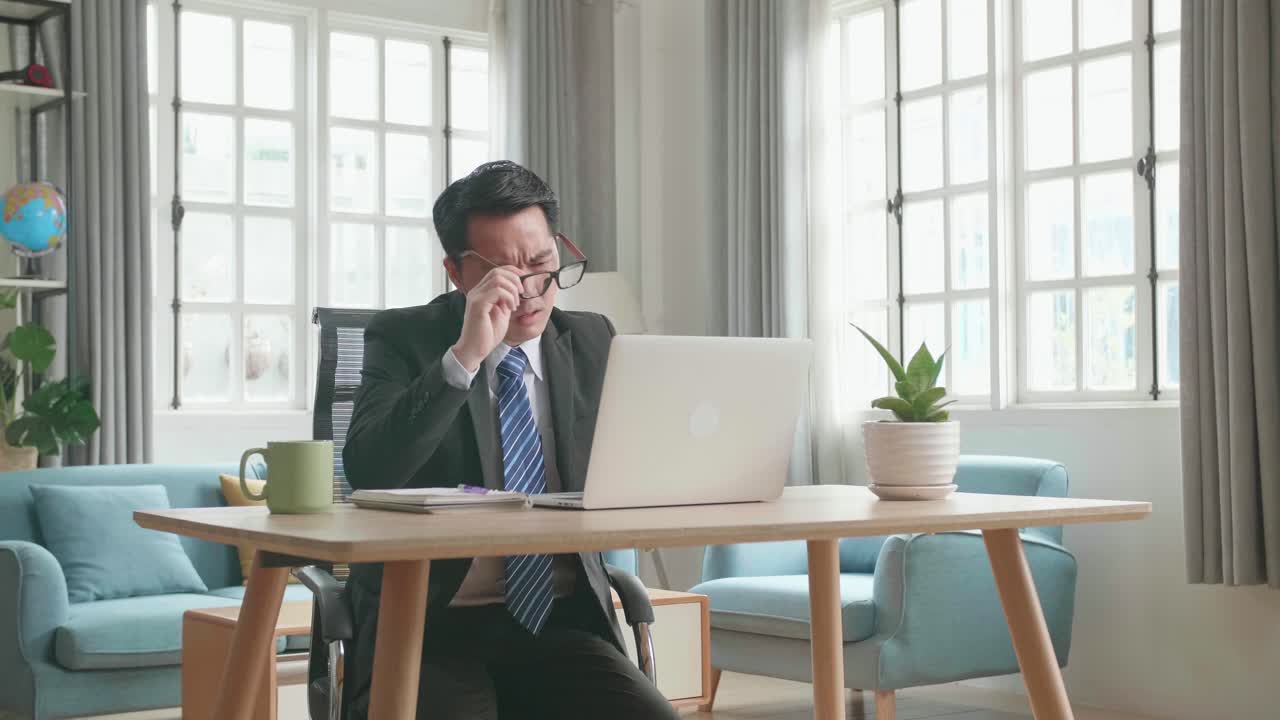 Tired Asian Businessman In Business Suit Taking Off The Glasses While Using The Computer For Working At Home.