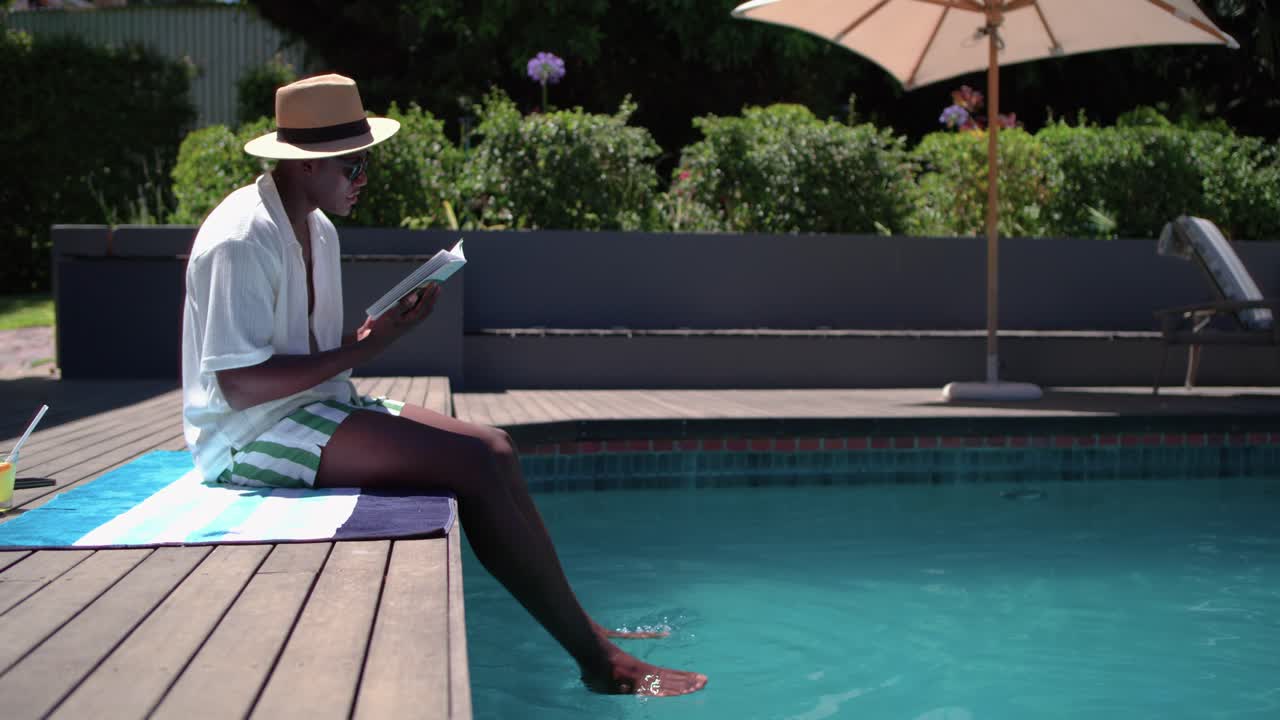 Man is settling at pool edge, reading paperback, sipping drink, dipping feet in water to relax