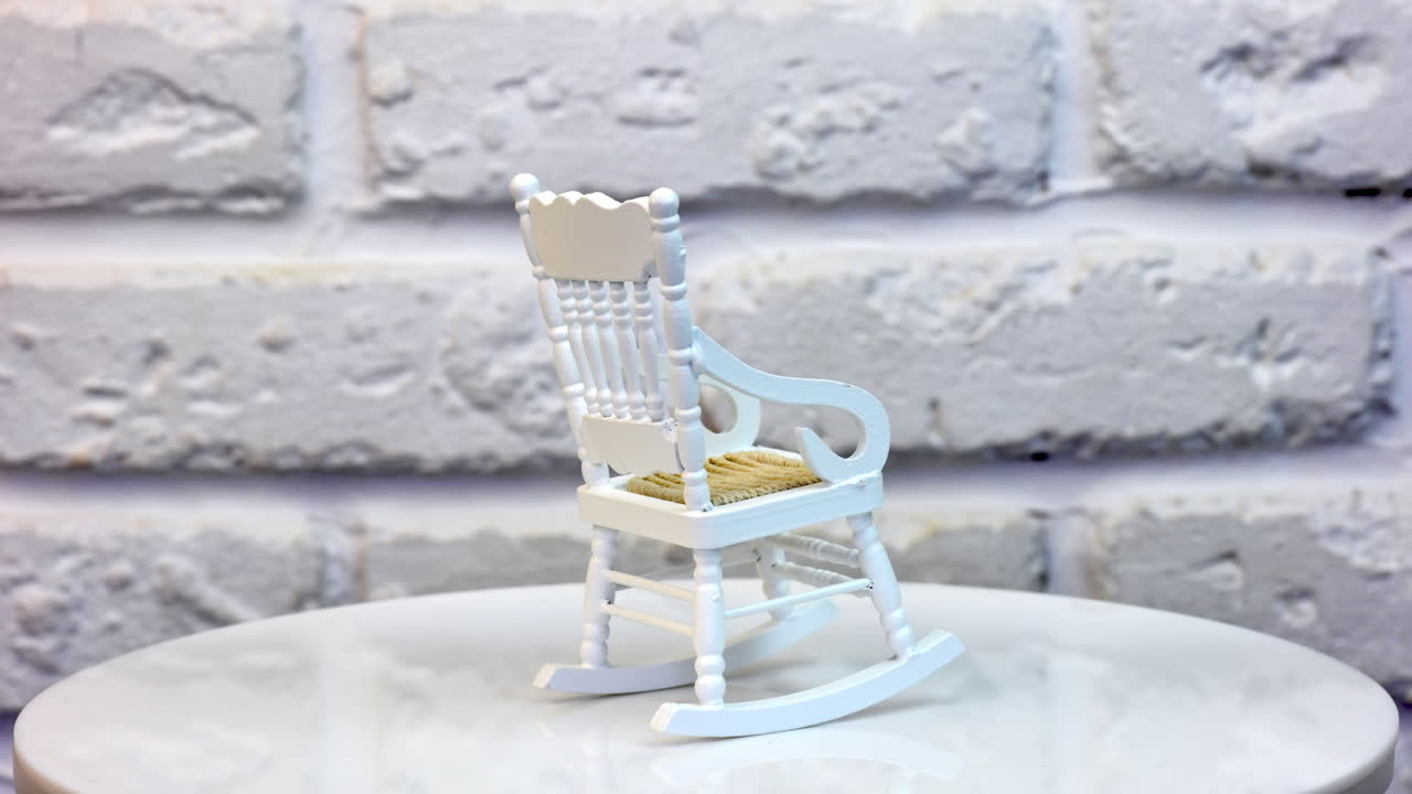 Little white rocking chair for doll's house. Pretty small piece of furniture on the rotating table. White brickwall at the background. Close up.