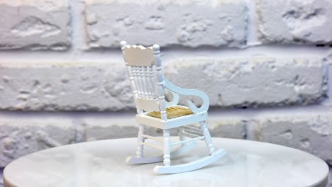 little white rocking chair for doll's house. pretty small piece of furniture on the rotating table. white brickwall at the background. close up.