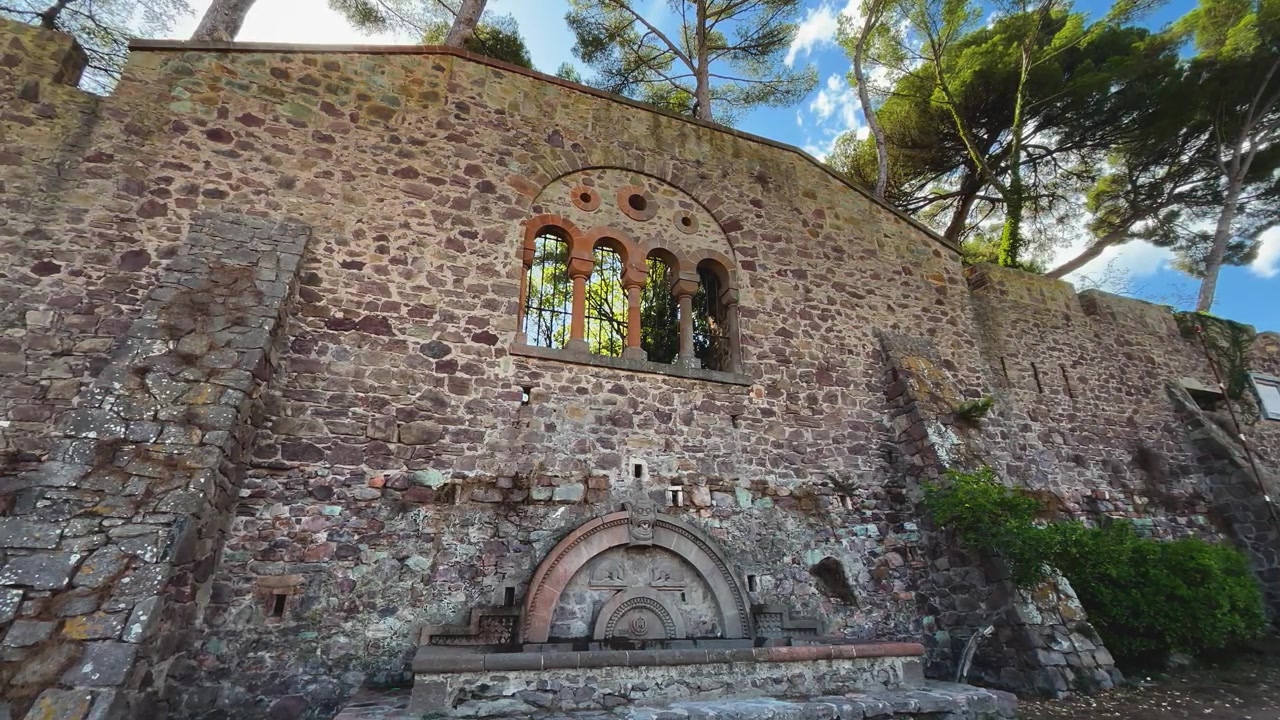 Un muro in pietra consumato presenta archi romanici, elementi in ferro battuto decorativi e una fontana intagliata e ornata sotto alti pini mediterranei.