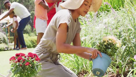 Madre-Afroamericana-Plantando-Flores-Arrodillada-En-Un-Jardín-Soleado,-Familia-En-El-Fondo,-Cámara-Lenta