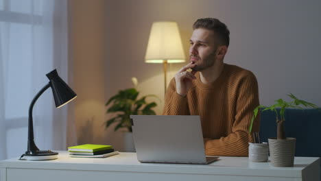 young guy is wearing brown sweater is sitting in front of table with laptop at evening working at home creative profession and remote work