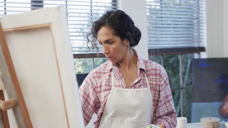 Focused-biracial-female-artist-in-apron-painting-on-canvas-in-studio,-slow-motion