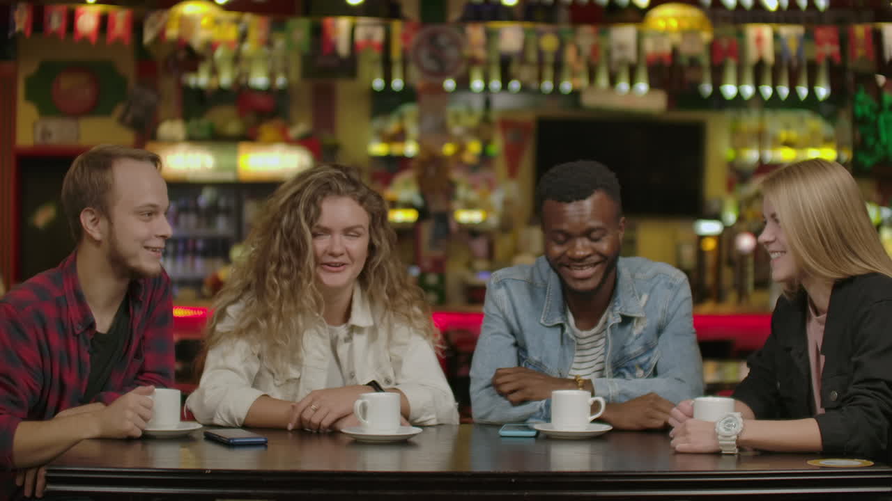 Bell'uomo ispanico mostra cose interessanti sul suo smartphone ai suoi amici mentre si divertono in un bar. Ridono, scherzano e bevono in un elegante bar hipster.