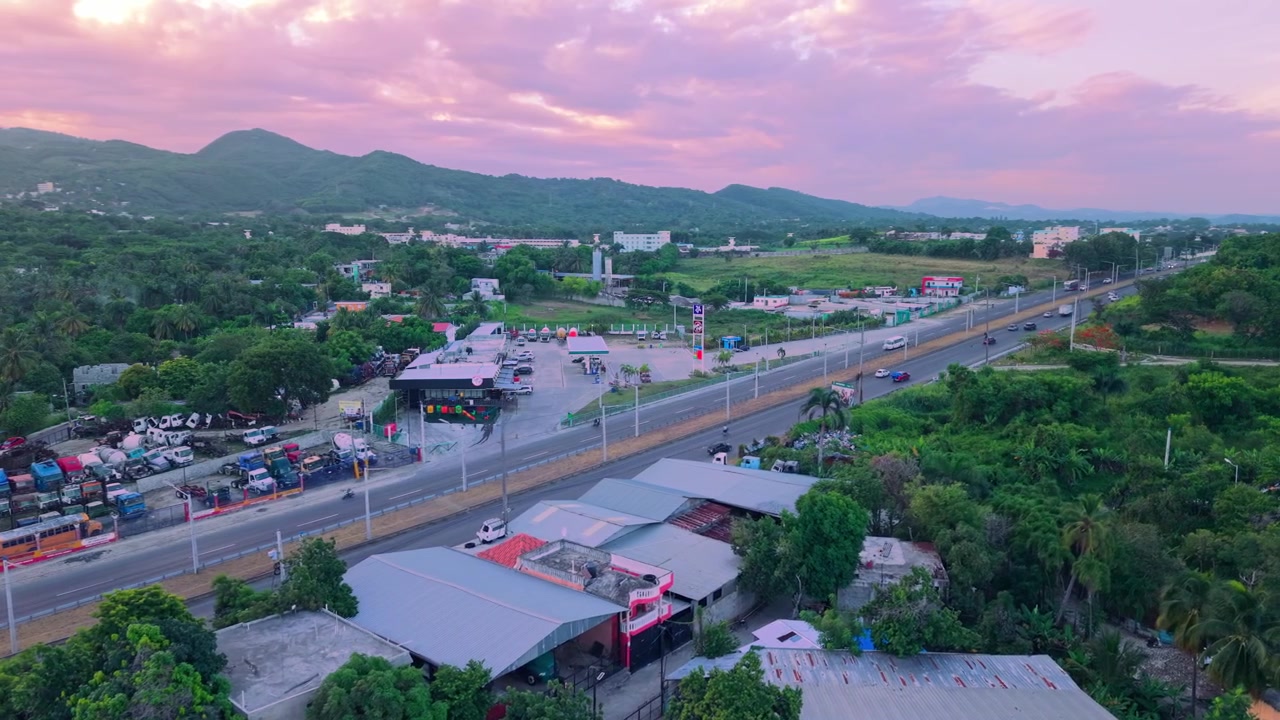 Imagem aérea de drone em ângulo elevado avançando sobre uma autoestrada de várias faixas ao pôr do sol, capturando o tráfego de veículos passando por um posto de gasolina, colinas verdes e um céu nublado rosado.