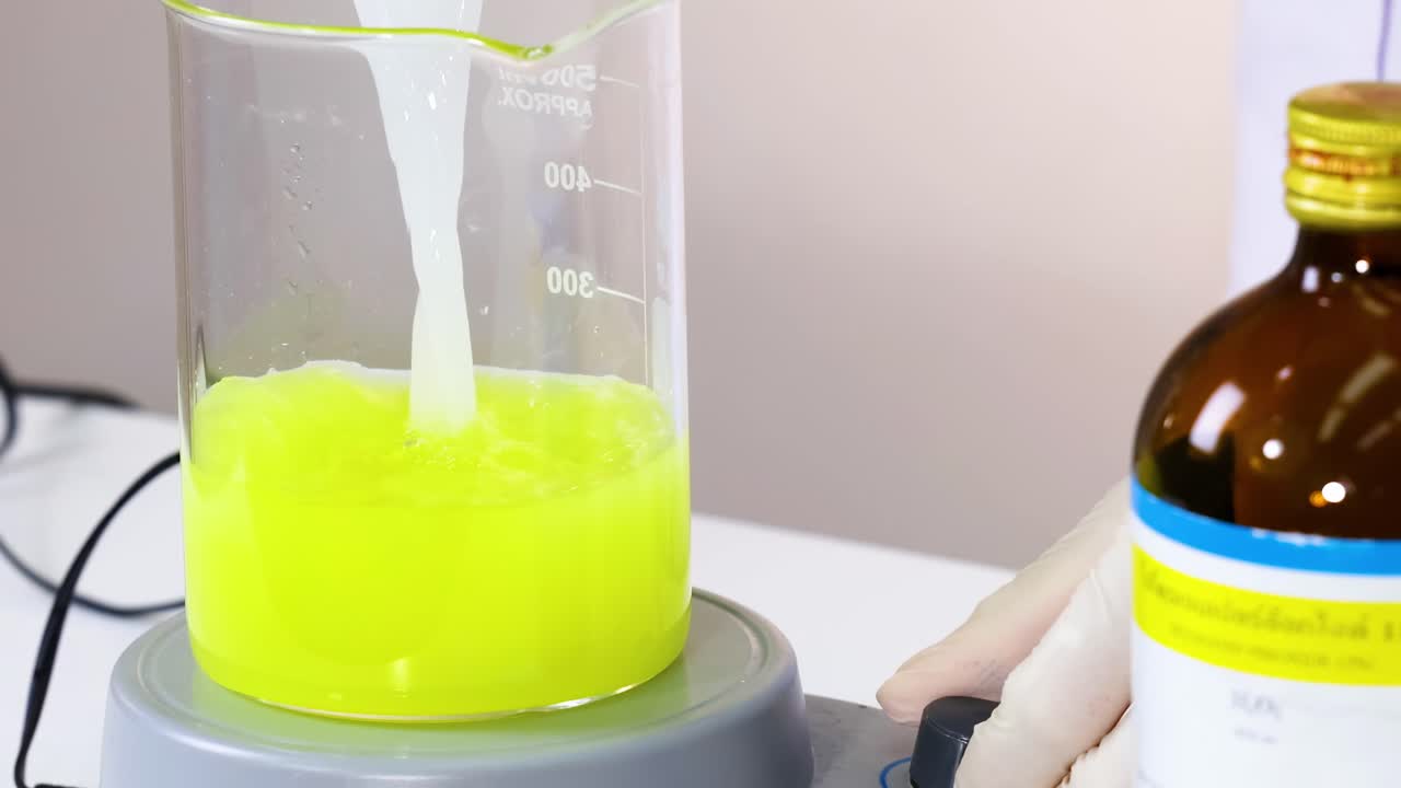 A vivid yellow liquid is stirred in a beaker using a magnetic stirrer, with a chemical bottle nearby.