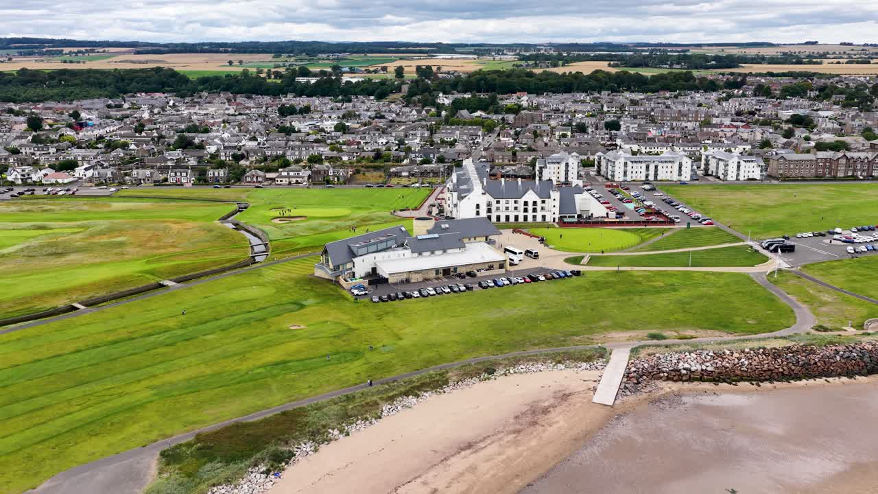 Drone pans across golf course, clubhouse, and town under cloudy daylight, revealing coastal landscape