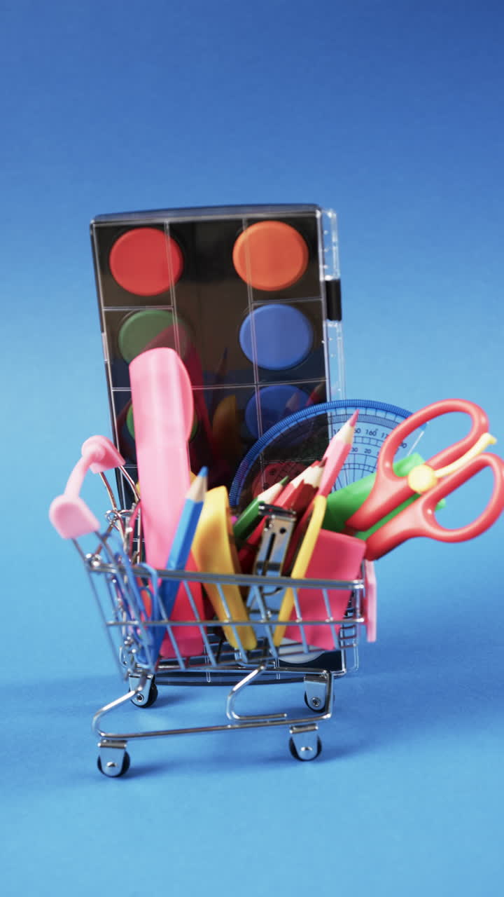 Vertical video of close up of shopping trolley with school items and paints on blue background