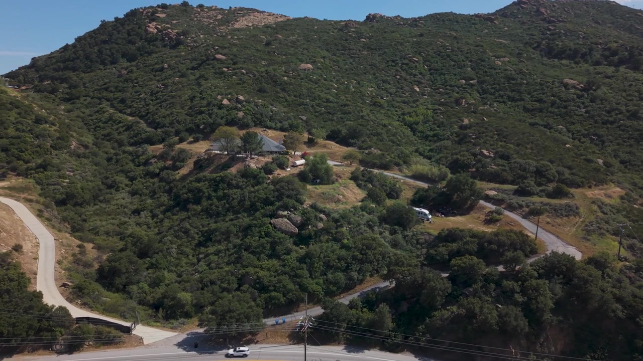 Lussuosa casa e villa in collina a Santa Susana Pass, California, circondata da un paesaggio di colline verdi con auto che percorrono una strada tortuosa in un suggestivo ambiente montano sotto un cielo blu. Ripresa aerea orbitale.
