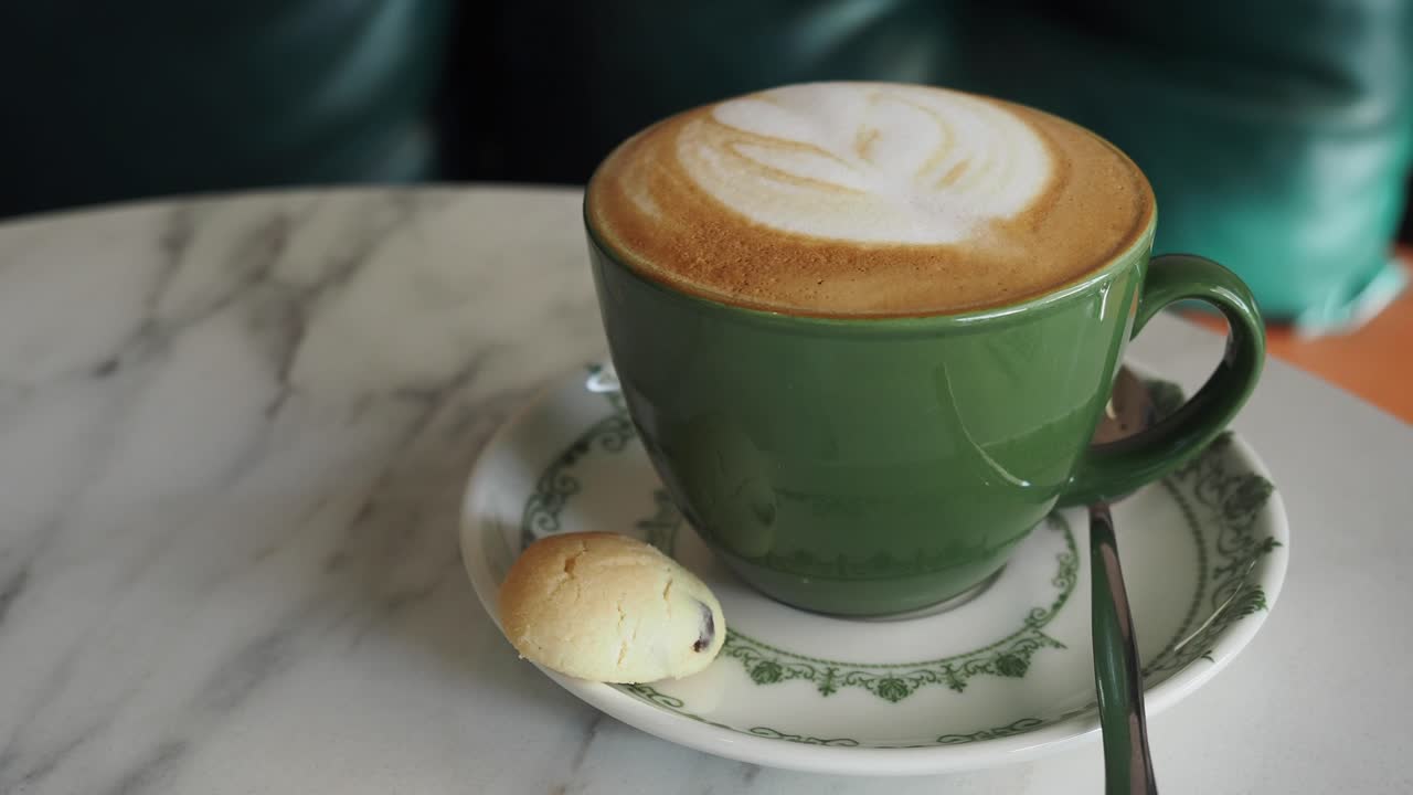 Closeup of a green coffee cup with a latte, a cookie, and a spoon on a marble table