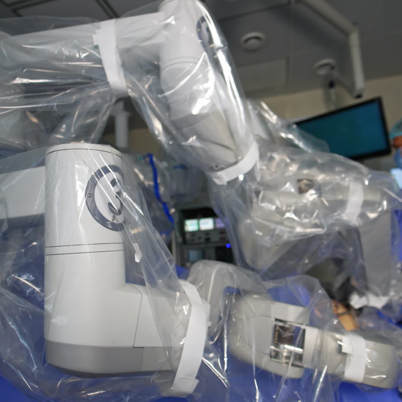 Massive robotic arms coated with plastic move at operation. Male surgeon stands behind the Da Vinci equipment