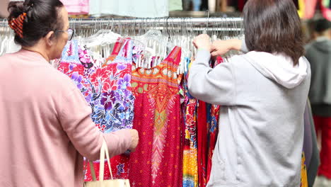 two women looking through a rack of colorful clothes at an outdoor market or garage sale in chinatown