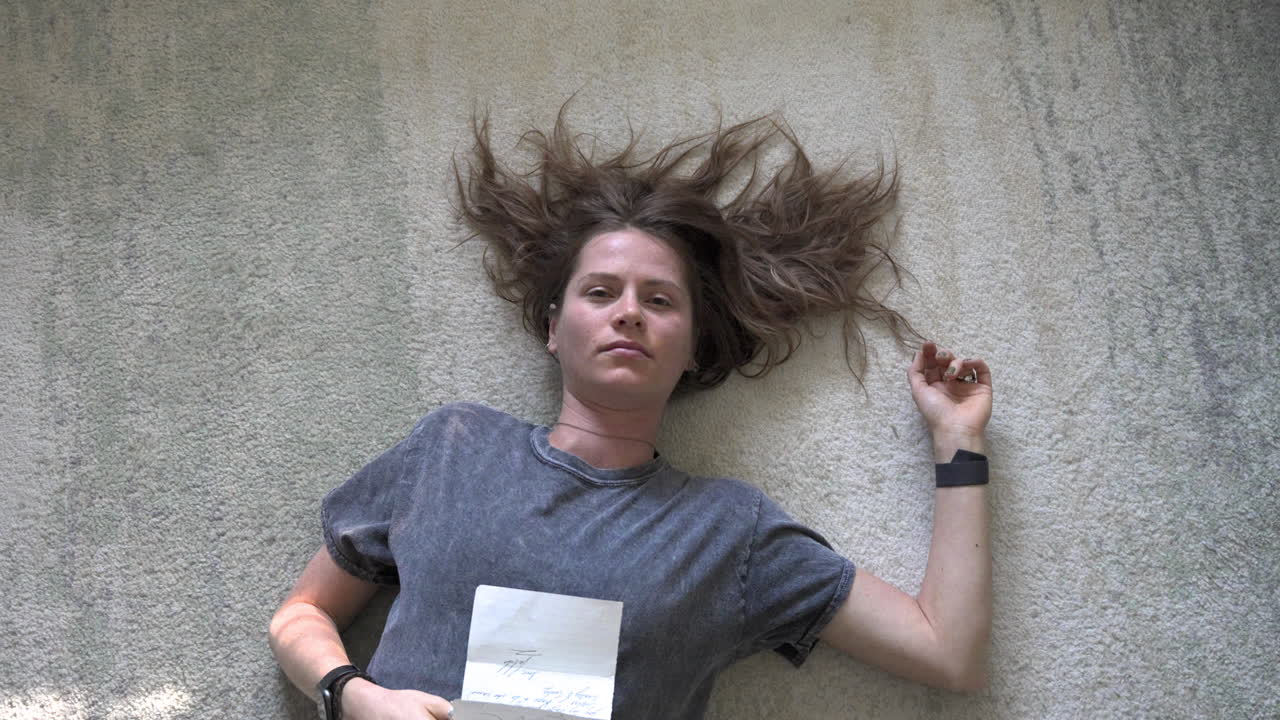 A serene topshot of a woman lying down with a love letter on her stomach, as the camera gently descends and rotates around her.