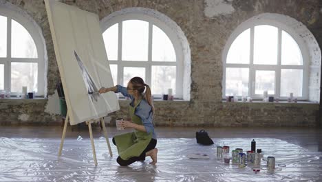 female artist working with large paintbrush and wide canvas in workshop