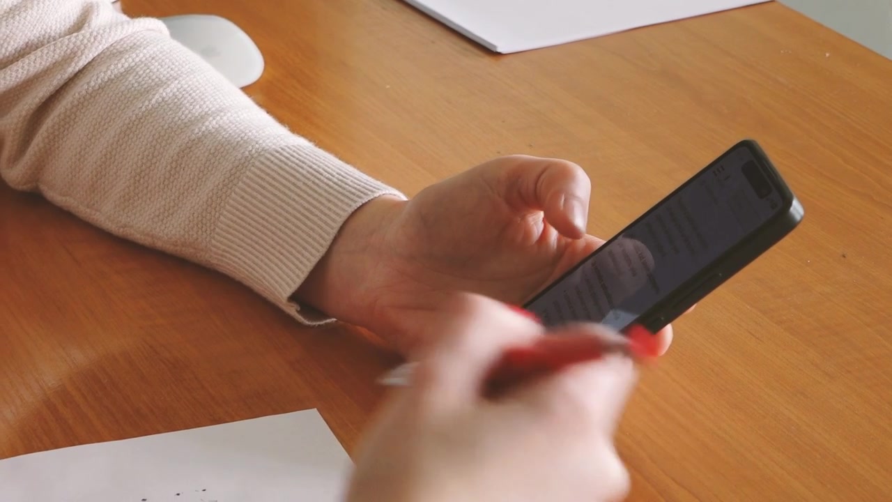 Una persona irriconoscibile tiene uno smartphone su una scrivania di legno con dei documenti nelle vicinanze, mentre esamina il testo sullo schermo in uno studio o spazio di lavoro con spazio per il testo