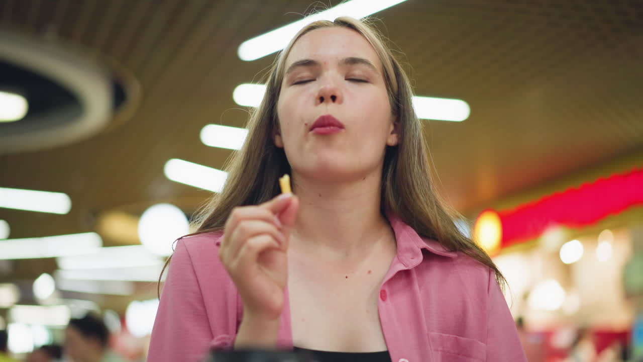 mujer sentada en un restaurante toma un bocado de papas fritas, cierra los ojos en satisfacción, y sacude la cabeza, disfrutando del delicioso sabor de la comida, luces brillantes del restaurante con fondo borroso