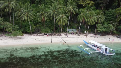 Tourist-people-of-an-Island-hopping-tour-boat-having-lunch-stop-at-Pasandigan-Cove-of-Cadlao-island-in-El-Nido,-amid-tropical-scenery-of-coconut-Palm-trees,-clear-water-and-white-sand-beach