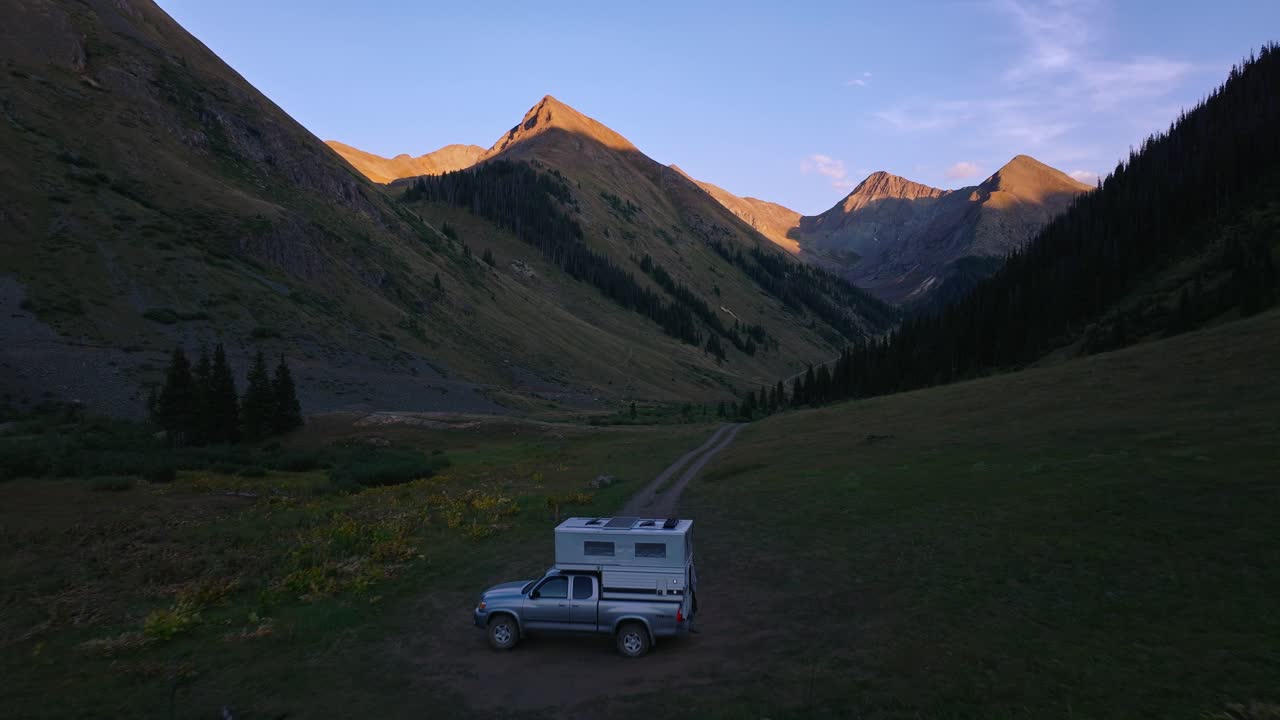 Una strada sterrata si snoda lungo una valle alpina erbosa sotto cime montuose risplendenti di bagliore alpino, ripresa aerea in allontanamento su un camper.