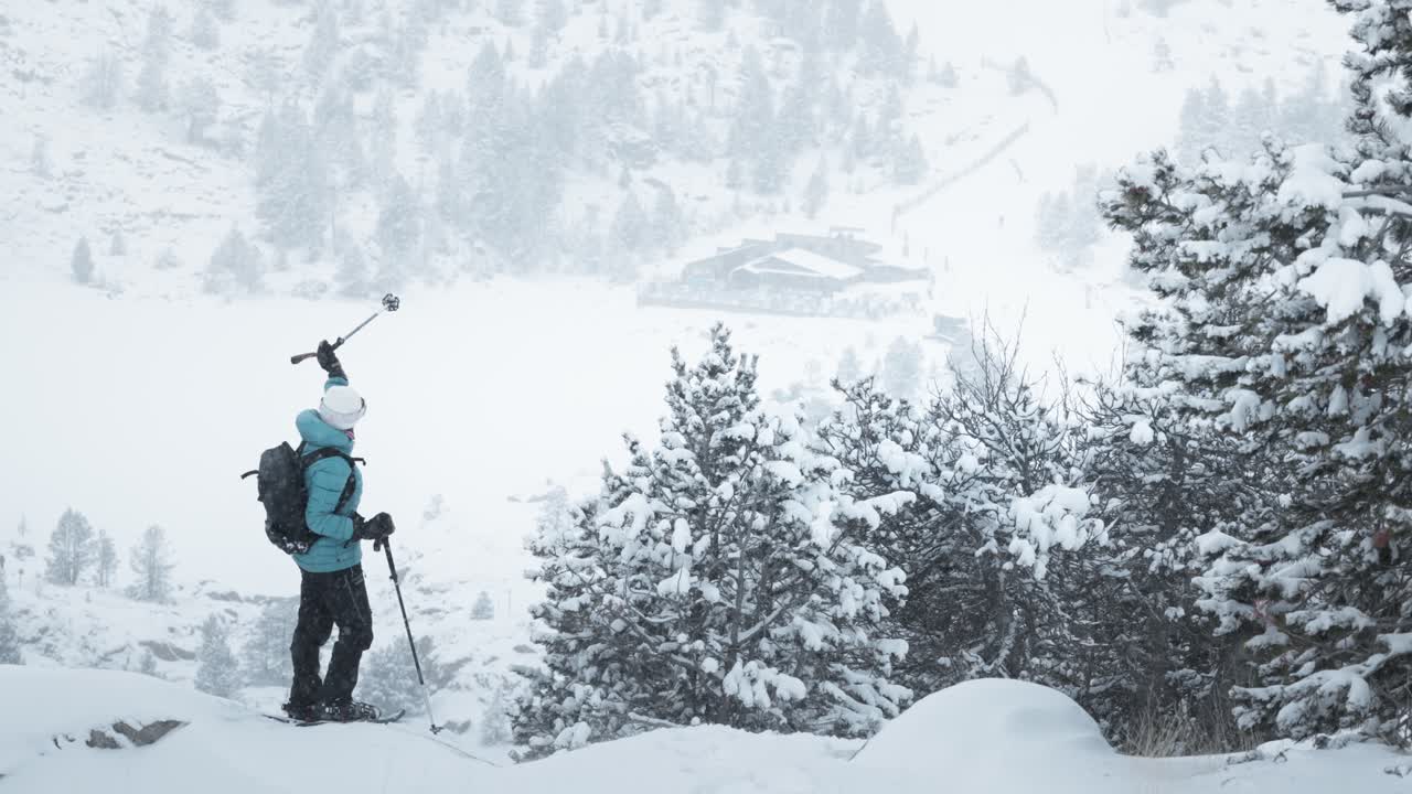 Static shot of a snowshoer pointing with a pole at snowy mountains, pine trees, and ski station in Andorra.