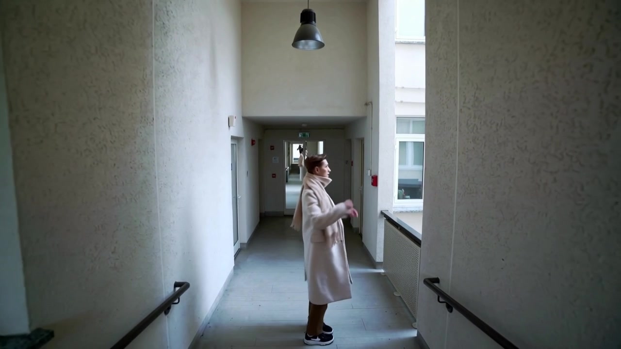 Avanzando, un uomo con un lungo cappotto passa accanto ai corrimano in un corridoio, sistemandosi la sciarpa verso l'uscita.