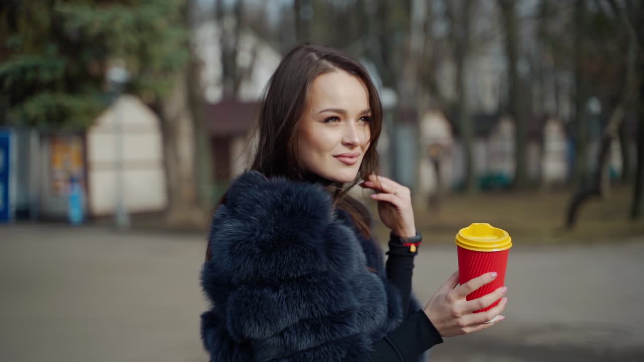 Beautiful woman with a plastic cup of coffee in the park. Portrait of brunette girl in fur coat posing on camera outdoors. Slow motion.