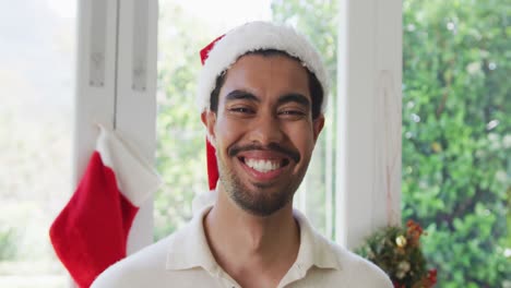 Portrait-of-happy-biracial-young-man-in-santa-hat-at-home-during-christmas