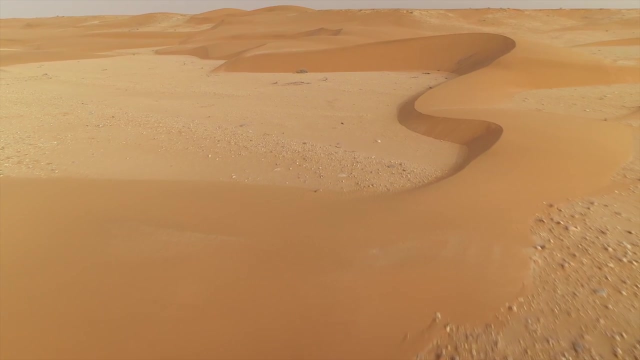 Volo di drone a bassa quota su terreno sabbioso piatto e arido verso dune desertiche ondulate a El Agunia, Marocco.
